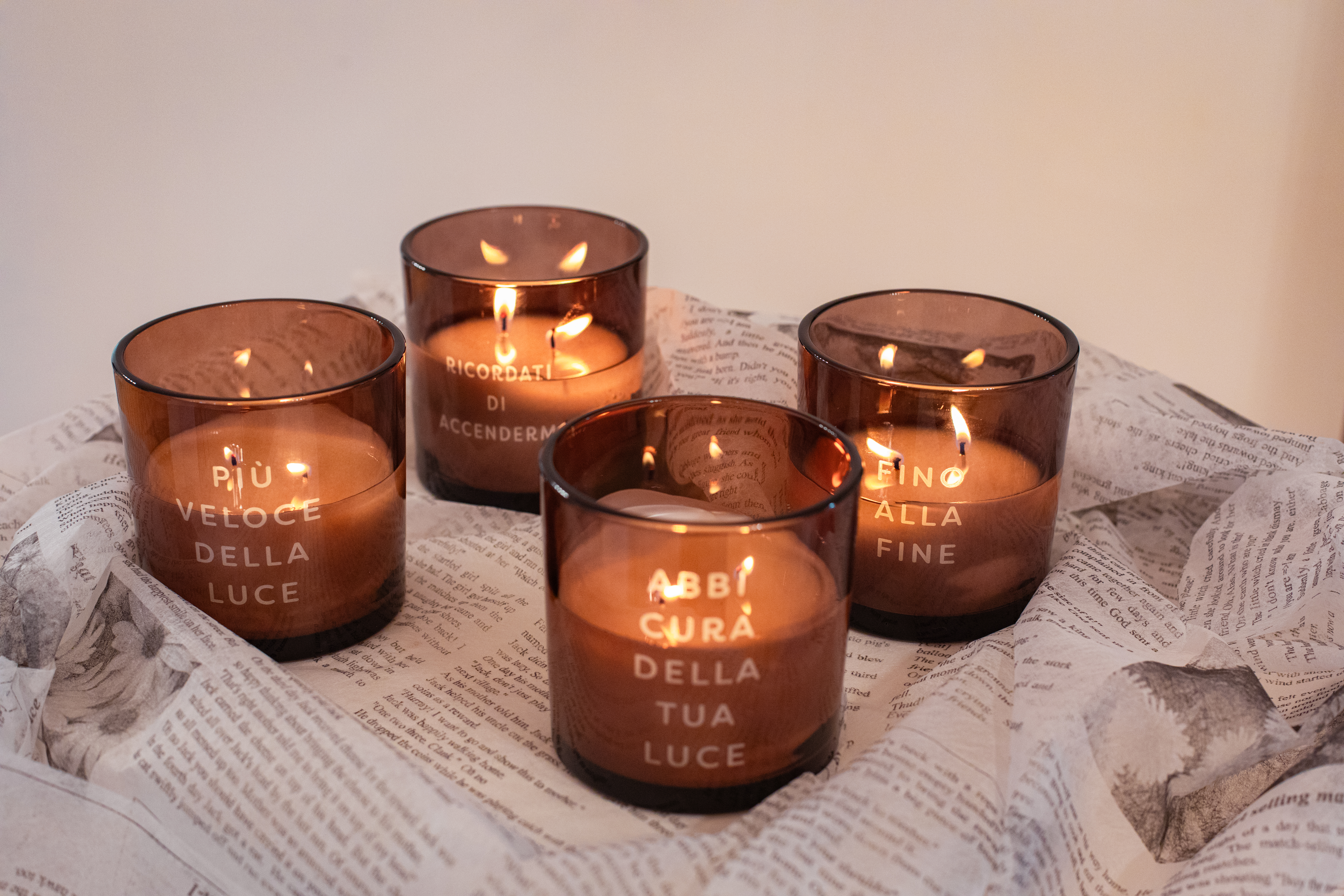 Quattro candele profumate della collezione Words & Lights di Pernici, in vaso di vetro ambrato con frasi incise: “Più veloce della luce”, “Ricordati di accendermi”, “Abbi cura della tua luce” e “Fino alla fine”, accese e disposte su fogli stampati che creano un’atmosfera calda ed elegante.